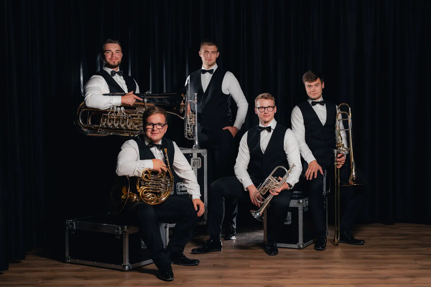 Silver Brass w białych koszulach i czarnych kamizelkach z instrumentami na tle ciemnej kurtyny